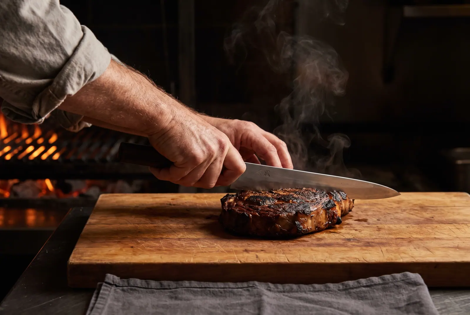 Hände tranchieren ein gegrilltes Steak auf einem Holzbrett, im Hintergrund die Glut